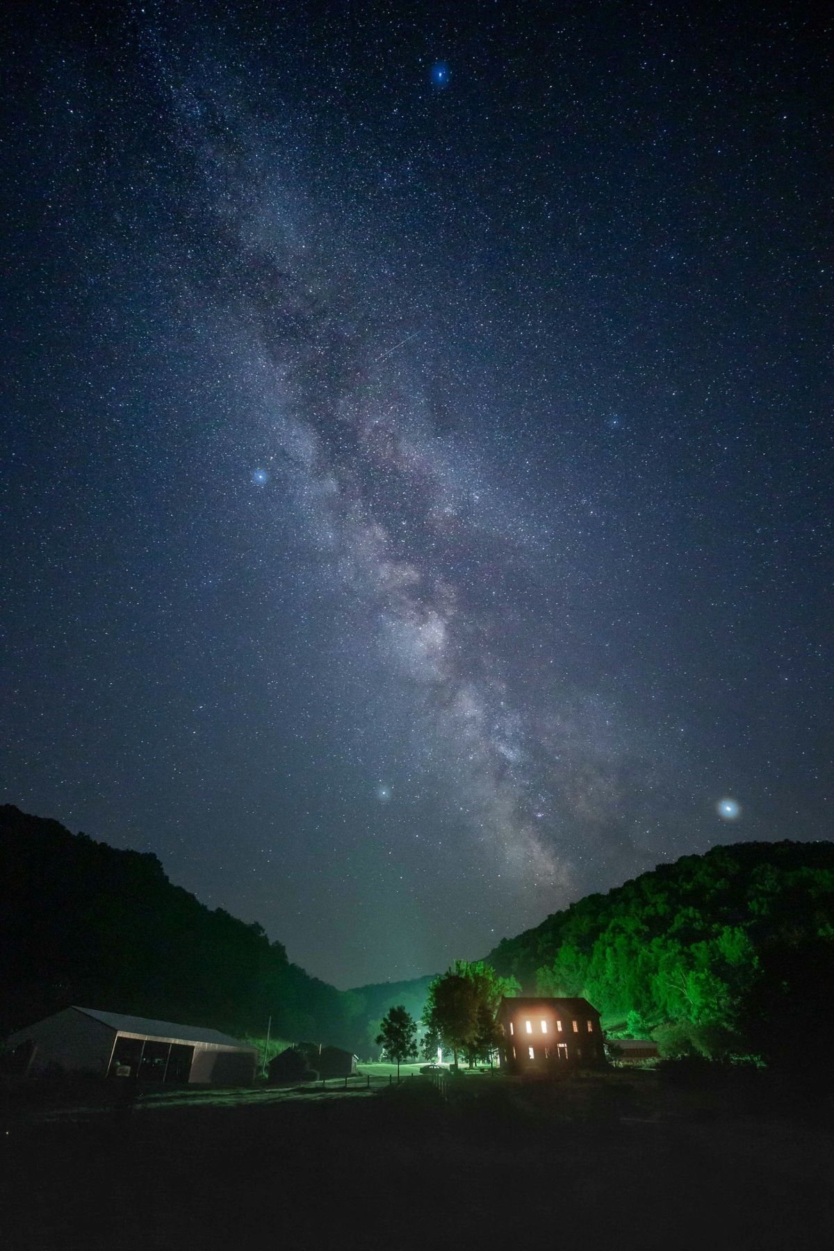 A farmhouse with its lights on under a night sky full of stars